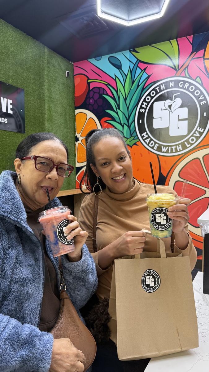 Two guests holding Smooth N Groove smoothies in front of the fruit mural